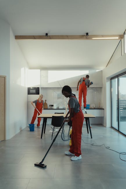 A spacious, modern kitchen area being professionally cleaned by three staff members from [COMPANY_NAME], engaged in surface cleaning and sanitisation. In the foreground, a woman using a vacuum cleaner
