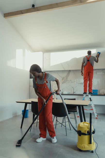 A spacious, modern kitchen area being professionally cleaned by three staff members from [COMPANY_NAME], engaged in surface cleaning and sanitisation. In the foreground, a woman using a vacuum cleaner